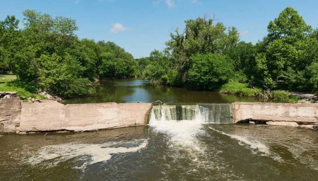 View of a small waterfall spilling over a concrete wall along the river in Kansas. You can see a wealth of green trees sitting along the edge of the river in the background. 