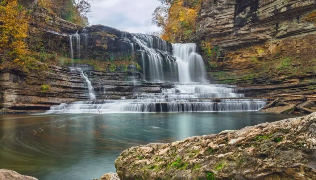 View of a small central waterfall tumbling down a series of rocks into a small blue pool. The waterfall is surrounded by rocks and vibrant fall foliage. 