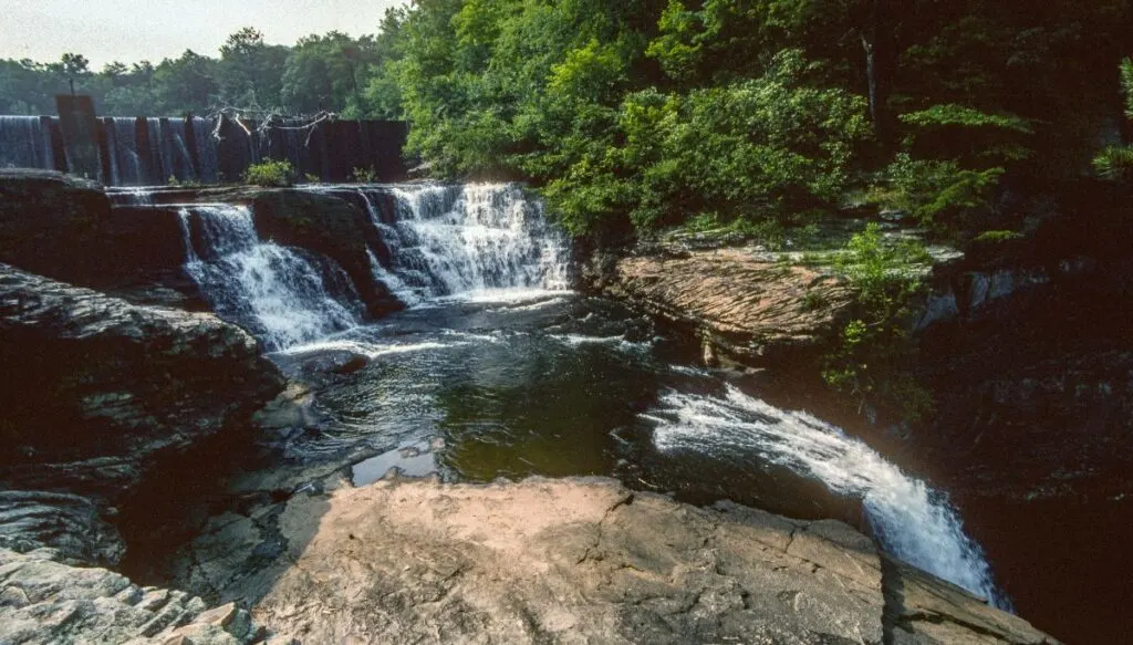View of two different waterfalls inside of DeSoto State Park. You can see a waterfall on the left that is surrounded by green trees on the right. 
