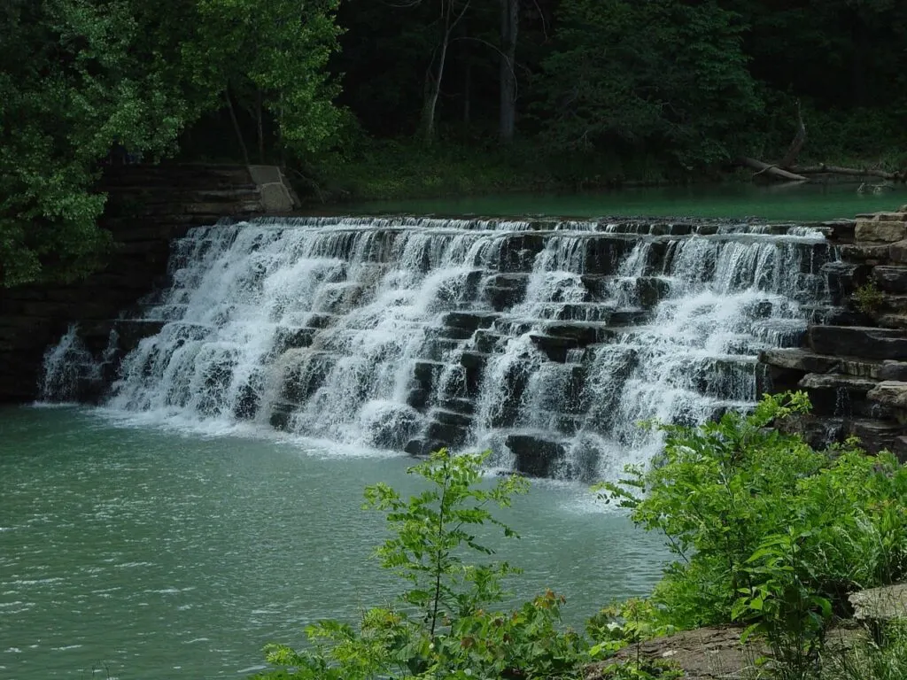 View of a broad waterfall cascading down into a river in Devil's Den State Park. The river above and below is light blue and surrounded by green trees. 