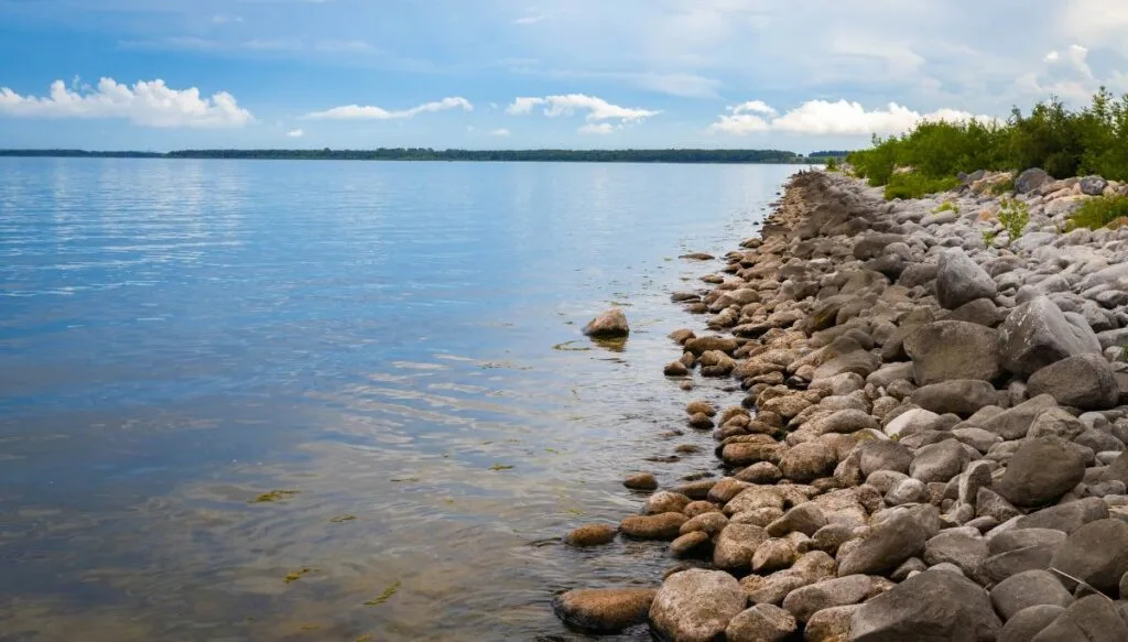 View of various gray stones sitting along the edge of Devils Lake with green land in the background on a cloudy day.