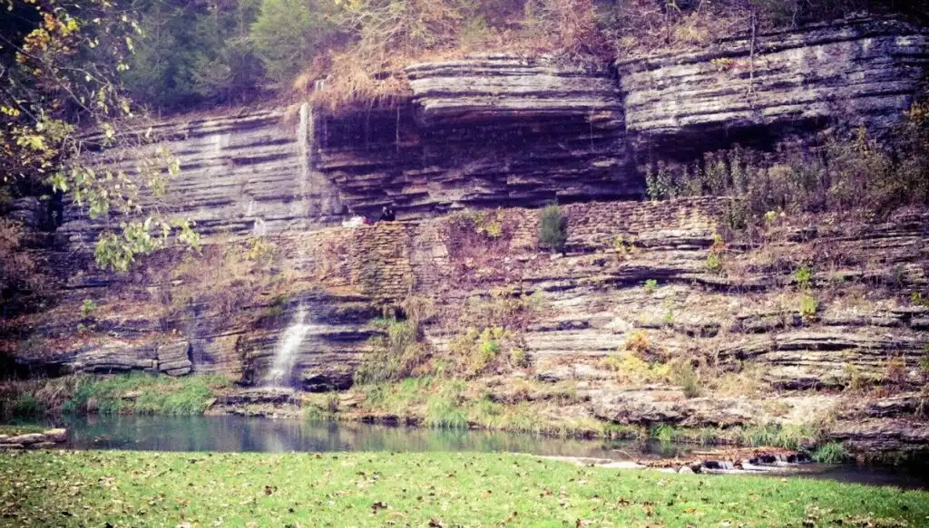 View of a waterfall falling down the side of a layered rock face with shrubs growing in the rocks and a pool at the base of the rocks that is surrounded by grass. 