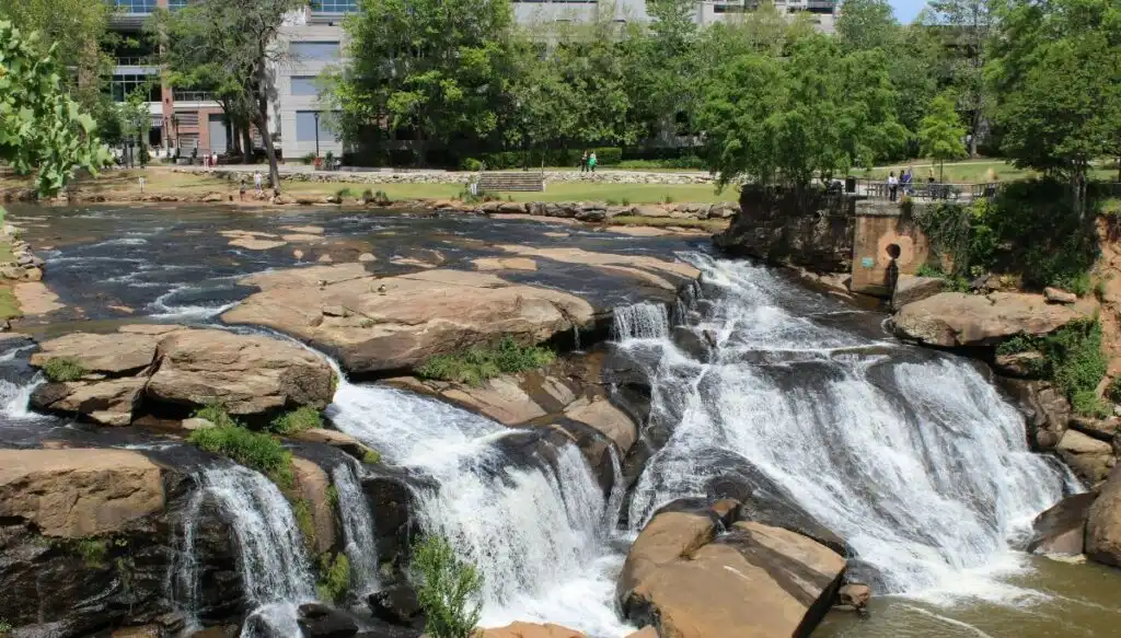 View of the large waterfall in the center of Falls Park on the Ready. You can see large rocks around the waterfalls and people standing at a viewing platform with trees and buildings in the background. 
