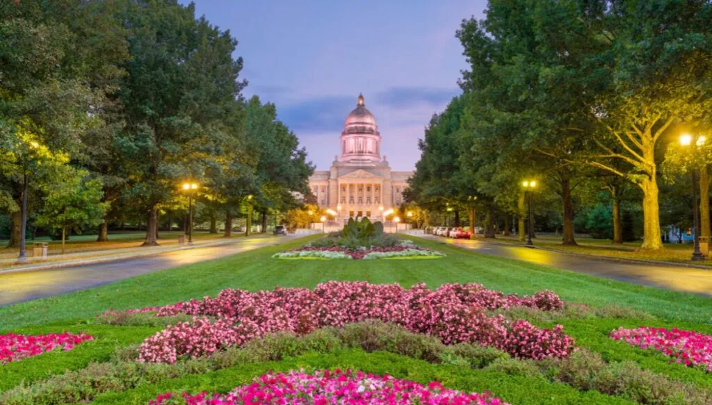 View of the state capitol building in the background and a beautiful garden with pink flowers in the foreground. You can see two walking paths on either side of the garden that are lined by green trees. 