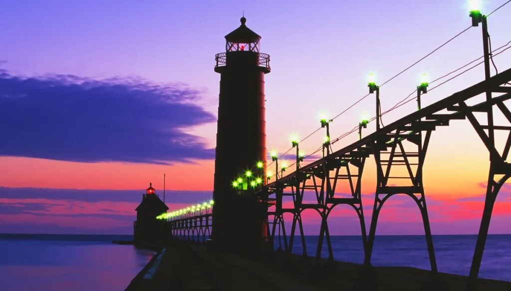 View of Grand Haven Lighthouse with a boardwalk in front of it and the sun setting in the background.
