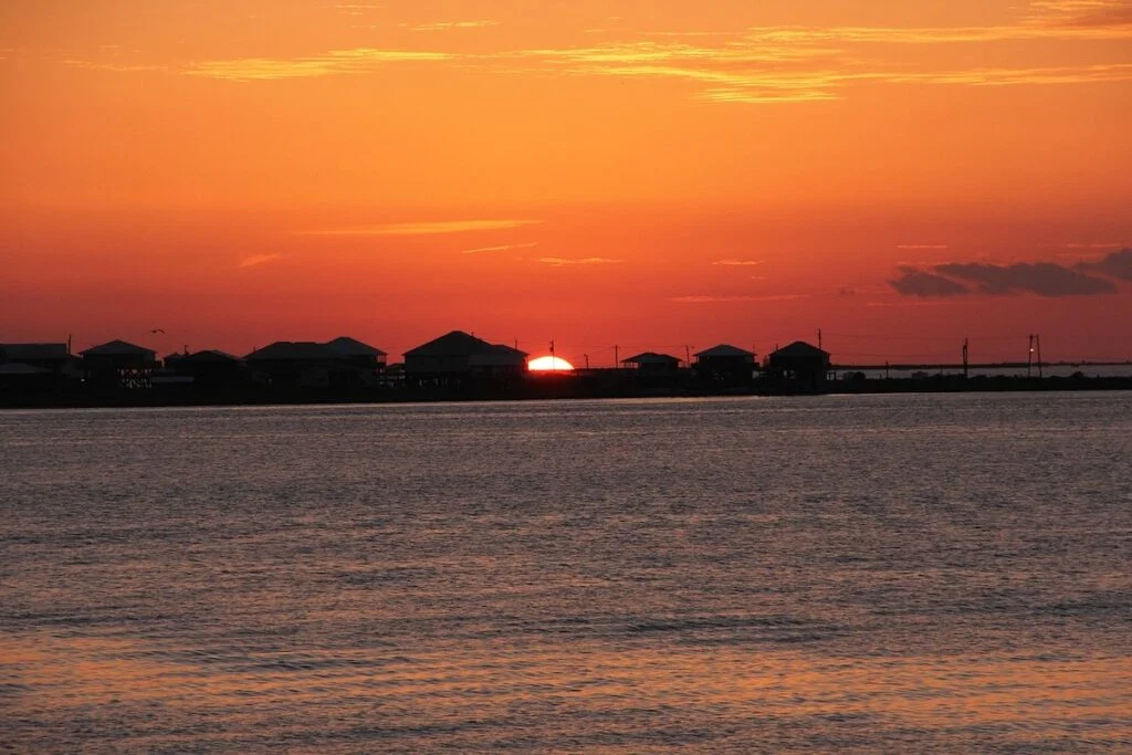 View of the sun setting behind houses with the water in front at Grand Isle State Park. 