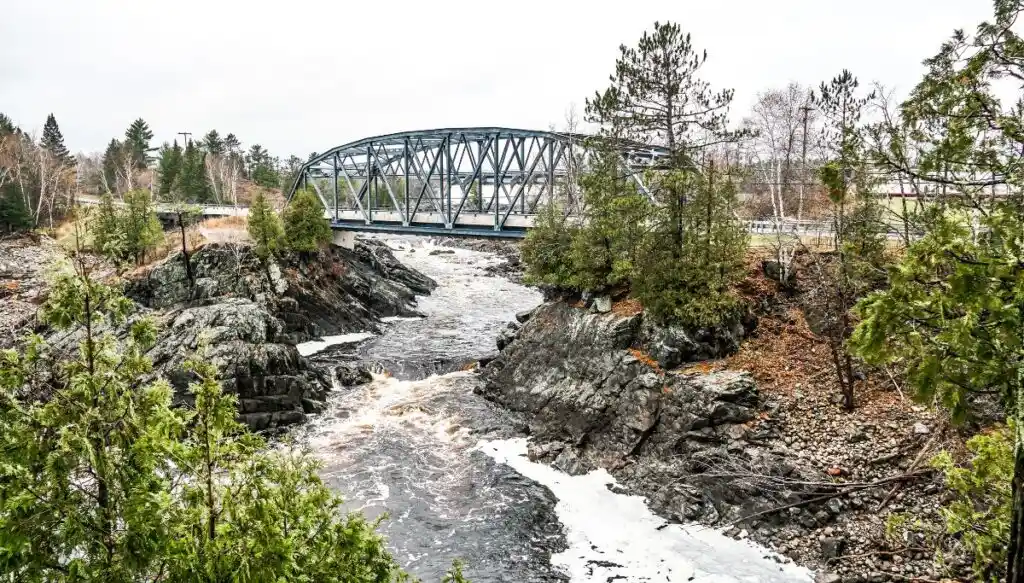 View of a metal bridge extending over a small river lined with rocks and pine trees. 