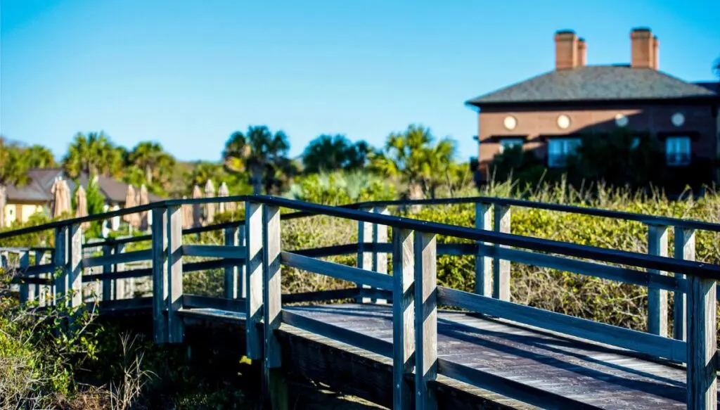 View of a wooden foot bridge surrounded by greenery with a brick house in the background. 