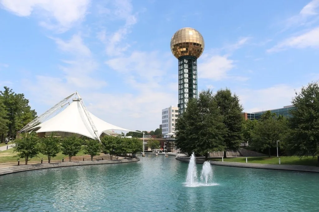 View of a tower with a gold ball on top that sits in front of a fountain with trees around it and a large white tarp hanging on the left. 