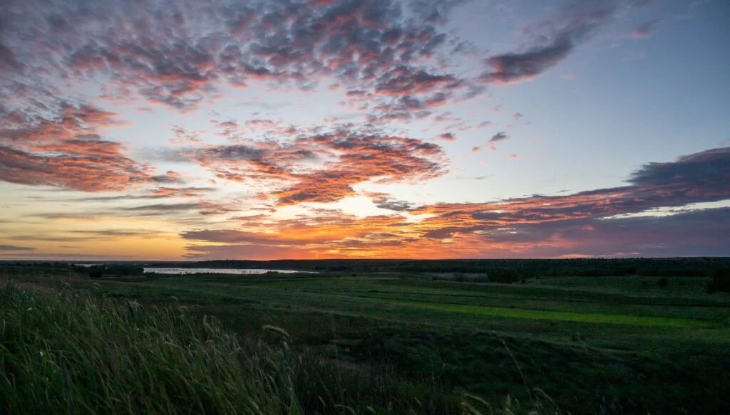 View of the sun setting over the vast green prairie in Kansas. It is dark and the sky has an orange color with the green prairie in the foreground.