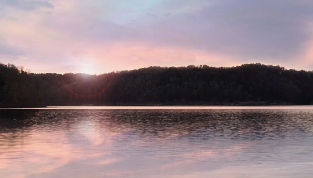 View of a lake surrounded by forests with the sun setting in the background and thw sky turning a a pale pink. 