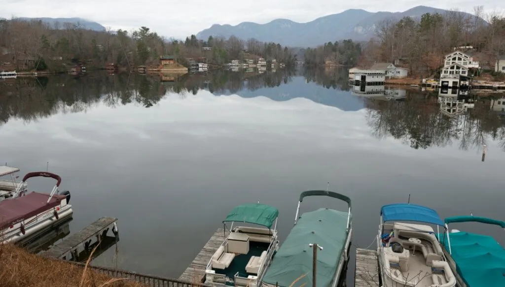 View of Lake lure in winter. You can see the bare trees around the lake and boats in the foreground docked at the pier. You can also see houses along the shore in the background. 