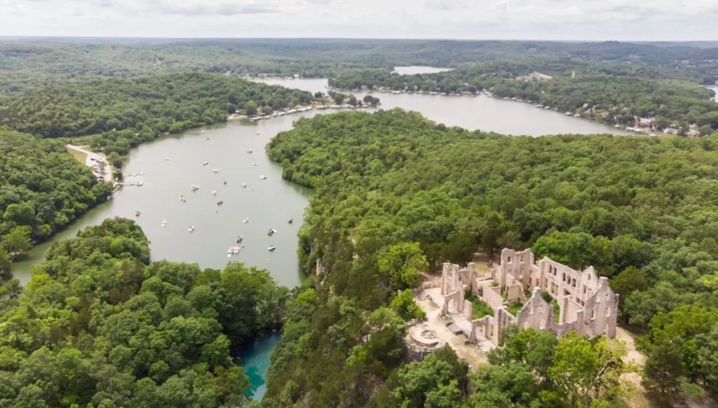 Aerial view of the Lake of the Ozarks. You can see the lake to the left and castle ruins to the right, all surrounded by vast forests of leafy green trees. 