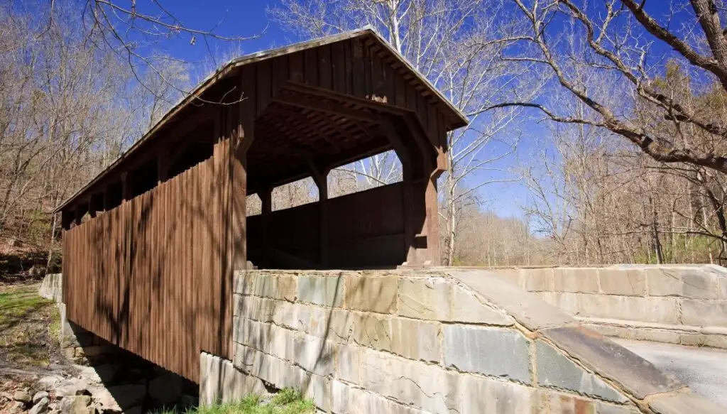 View of a brown, wooden covered bridge over a river with a stone wall in the front. 