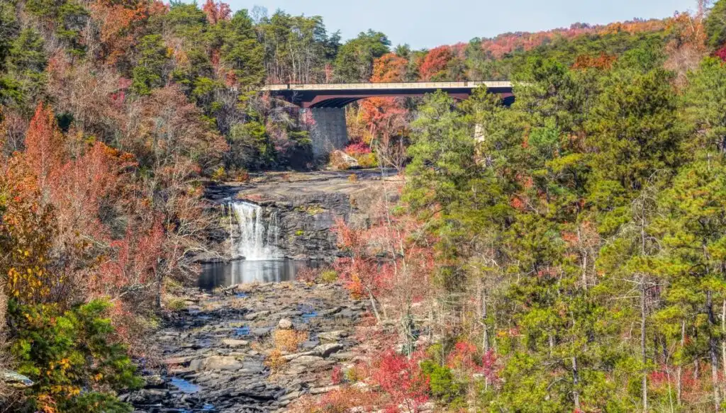 View of a bridge going across a river with a little waterfall in front that is surrounded by fall foliage. 
