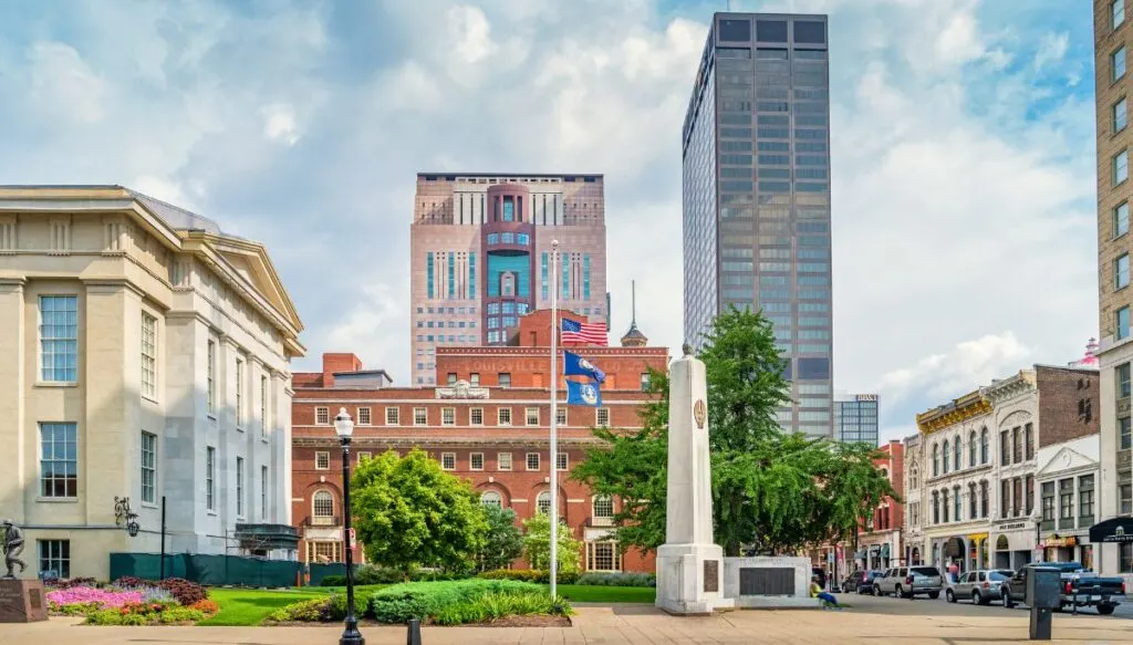 View of a historic brick building with a park in front of it and a white obelisk in downtown Louisville. 