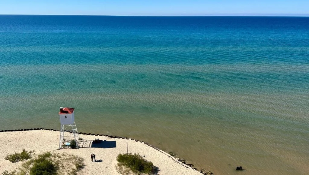 Aerial view of a beach with a white tower along the shores of a lake in Ludington State Park. You can also see two people walking along the beach towards the lake. 