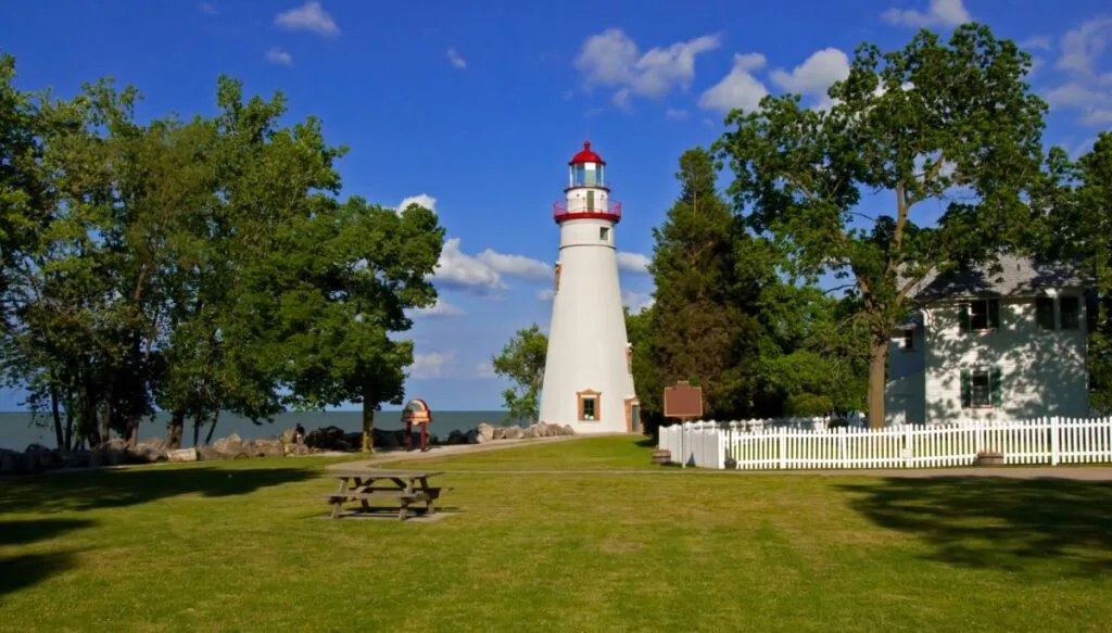 View of the red and white Marblehead Lighthouse in Ohio. It is one of hte most beautiful places in Ohio with a blue sky in the back and green trees on either side. 