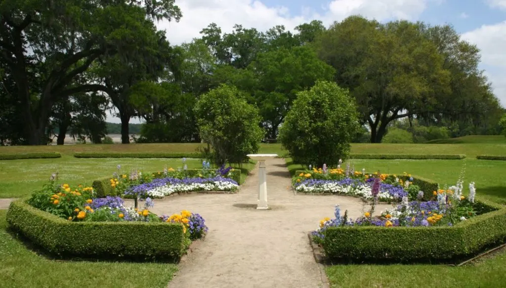 View of a beautiful garden with well-manicured flowers and hedges surrounding a small white bird path with walking paths and large green trees around it. 