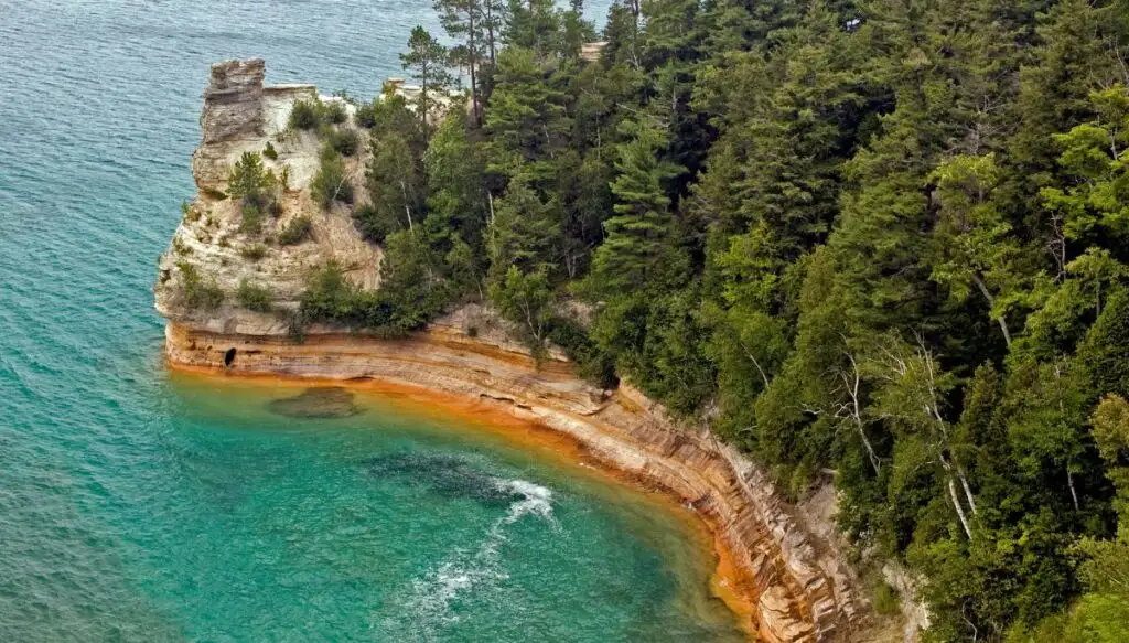 View of the ruins of a Miner's Castle along the edge of a lake shore filled with steep rocks and forests of pine trees. You can see birght blue water at the base of the lookout. 