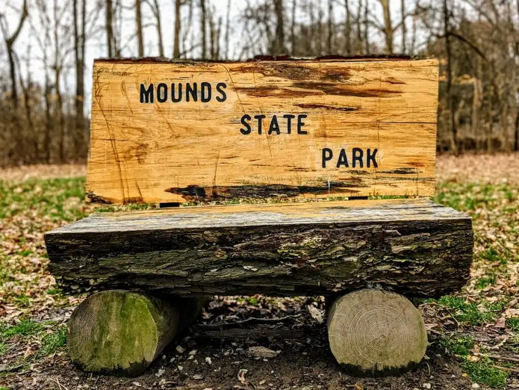 View of a bench made of whole logs that is very rustic. The back of the bench says, "Mounds State Park" in black letters and the bench is surrounded by forests. 