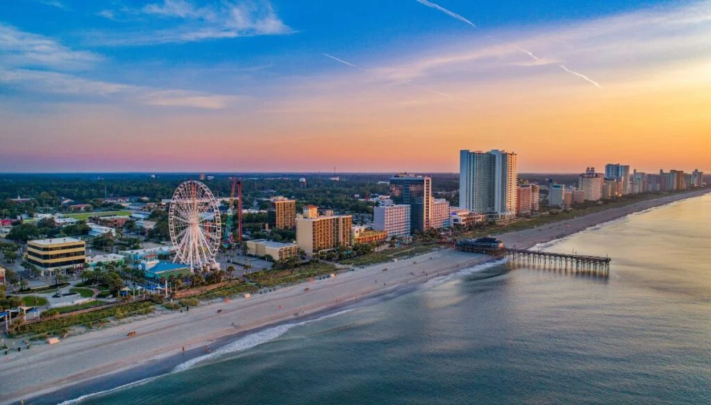 Aerial view of Myrtle Beach with a ferris Wheel and lots of tall buildings lining the beach as the sun sets in the background. 