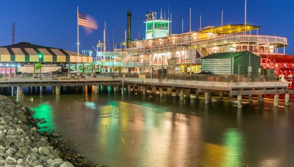 View of a river boat sitting in the water all lit up at night with a pier in front of it and an American flag to the left. 