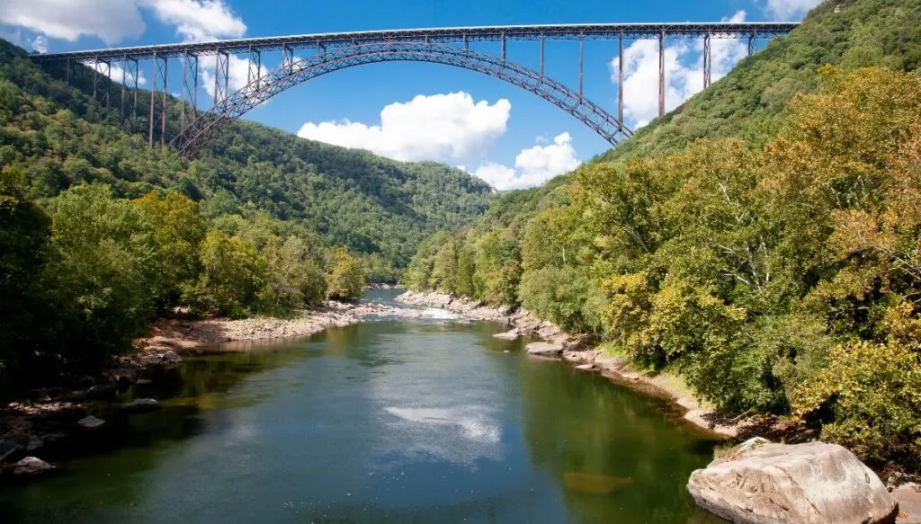 View of a metal bridge spanning across New River Gorge. You can see a river in the gorge that is surrounded by green trees with rocks along the shore of the river. 