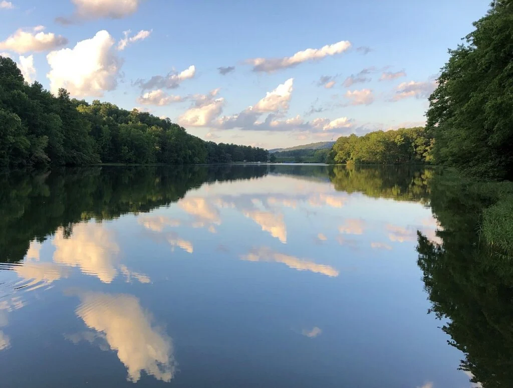 View of a lake in Oak Mountain State Park that is surrounded by green trees and that perfectly reflects the clouds on the surface of the lake. 