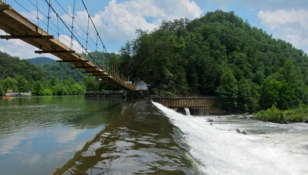 View of a walking bridge going across the Ocoee River Gorge with a dam in the middle and green trees in the background. 
