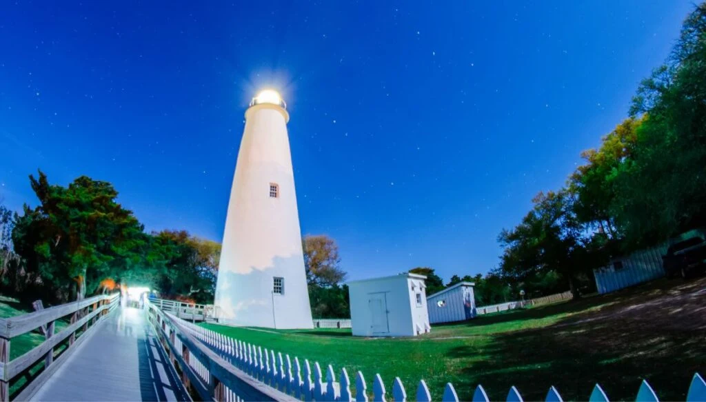View of the tall white Ocracoke lighthouse glowing atnight at the end of a white walkway. 