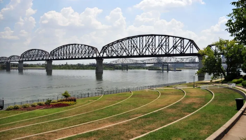 View of a metal bridge over the Ohio River. You can also see well-maintained gardens and grassy areas along the Ohio River in the foreground. 