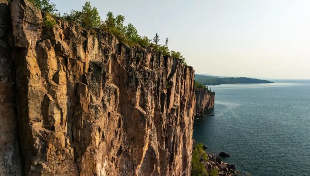 View of the sheer cliffs of Palisade Head with pine trees on top sna dthe blue waters of Lake Superior below. 