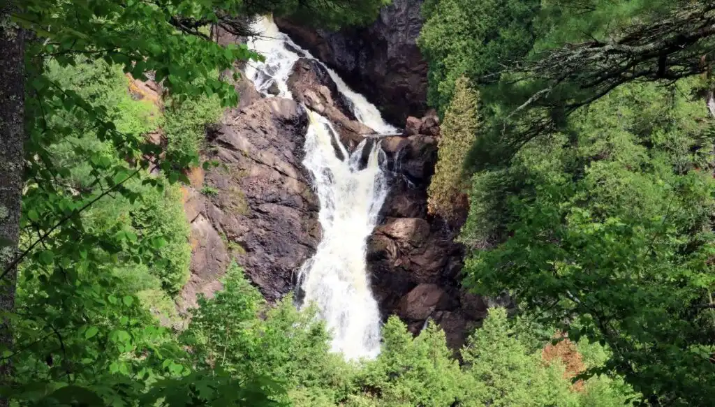 View of a large waterfall cascading down a series of rocks that are surrounded by green trees inside Pattison State Park. 