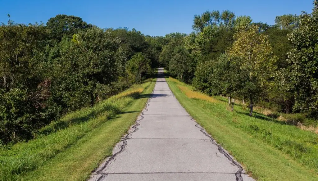 View of a paved walkway going through a green field in Pella, Iowa. You can see green trees all around the path. 