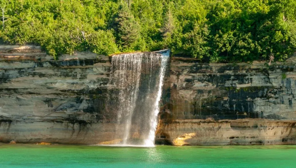 View of a small waterfall tumbling over the sheer edge of a cliff with tons of pine trees on top of the cliff and bright blue water below. 