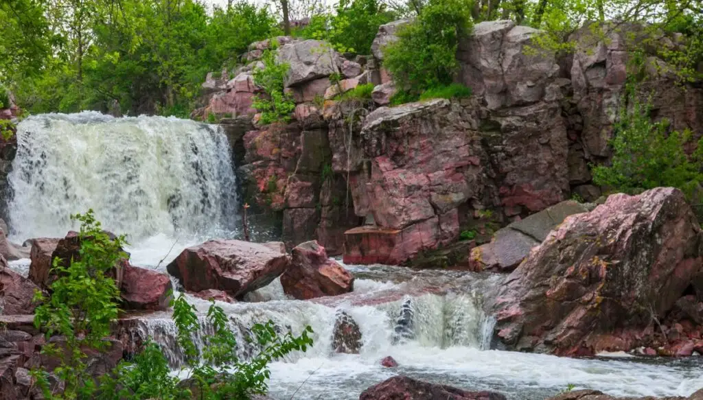 View of a small waterfall crashing down around large brown boulders with green trees all around it. 
