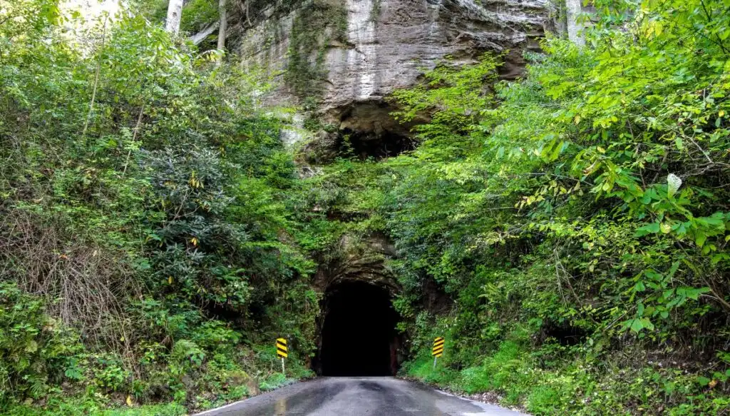 View of the haunted Nada Tunnel from the middle of a road in Red River Gorge. You can see rocks in the background and greenery all around the tunnel. 