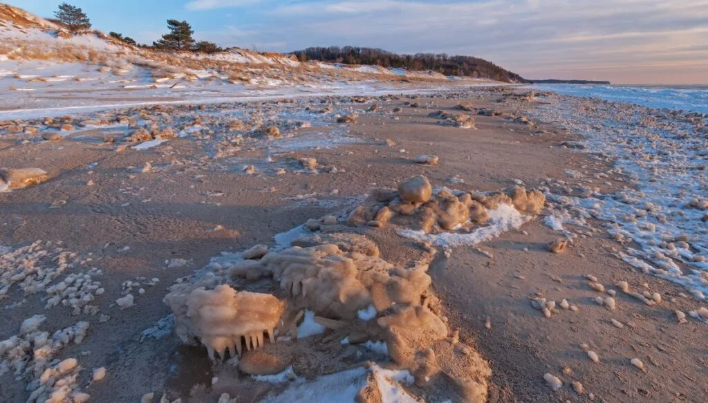 View of light snow dusting the sandy shores of Saugatuck Dunes State Park with the lake to the right and trees in the distance at sunset. 