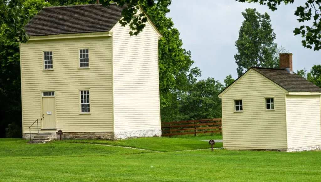 View of two simple white buildings from the Shaker Village in Pleasant Hill, Kentucky. You can also see tall green trees behind the buildings. 