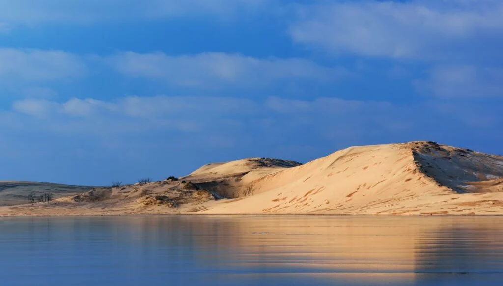 View of sand dunes sitting along the edge of a lake with a blue sky in the background. 