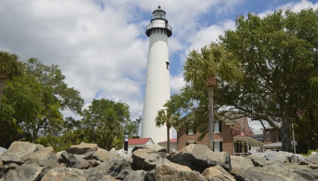 View of a giant white lighthouse tower with a wide variety of rocks sitting in front with green trees all around it. 
