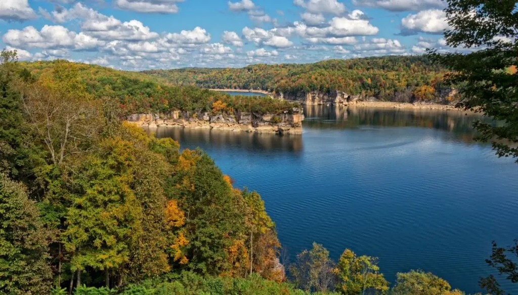 Aerial view of the blue waters of Summersville Lake. You can see trees slowly starting to change color along the edge of the lake and clouds in the air. 