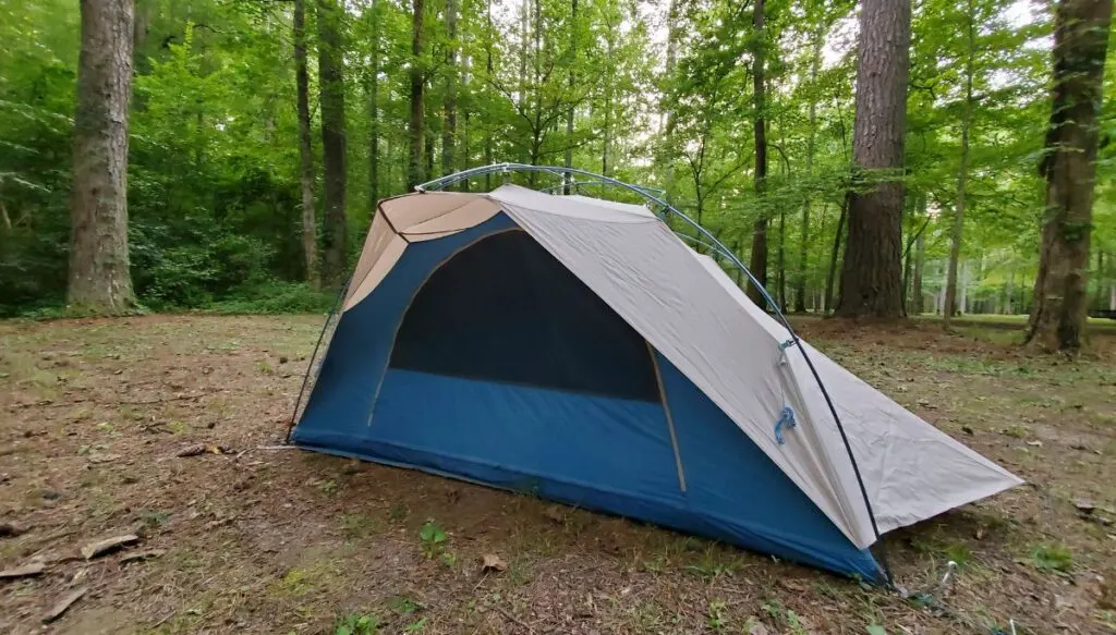 View of a blue and white tent sitting in a clearing in the middle of a forest surrounded by leafy green trees. 