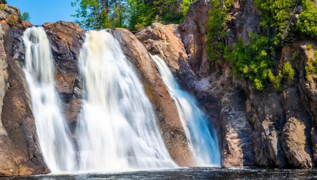 View of the massive high falls cascading down a series of rocks surrounded by pine trees in Tettegouche State Park.