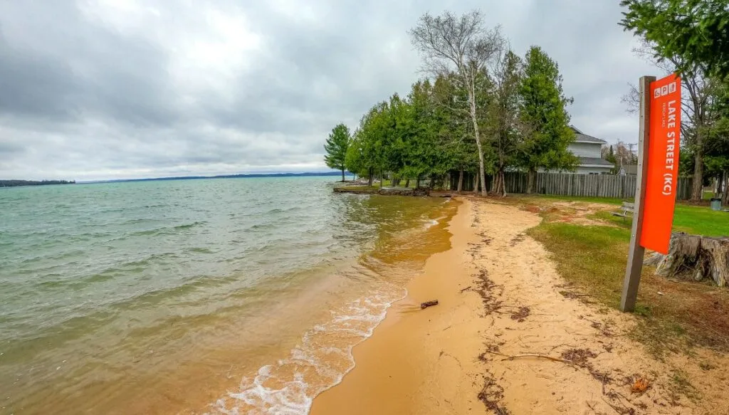 View of sandy beach along the edge of Torch lkae with pine trees in the back and a red sign in the grass on the right. 