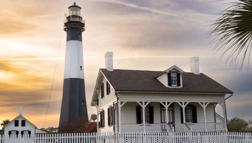 View of a small white cottage standing next to a tall black and white lighthouse with the sun setting in the background. 