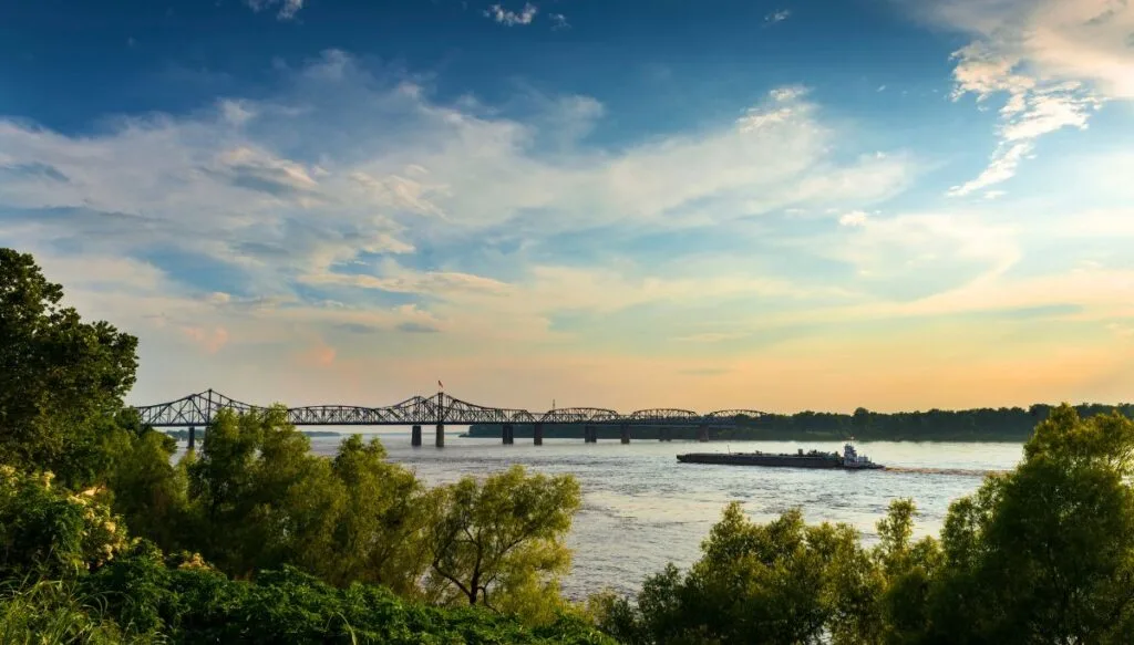 View of a bridge in the distance with boats going through the water and the sun setting in background. 