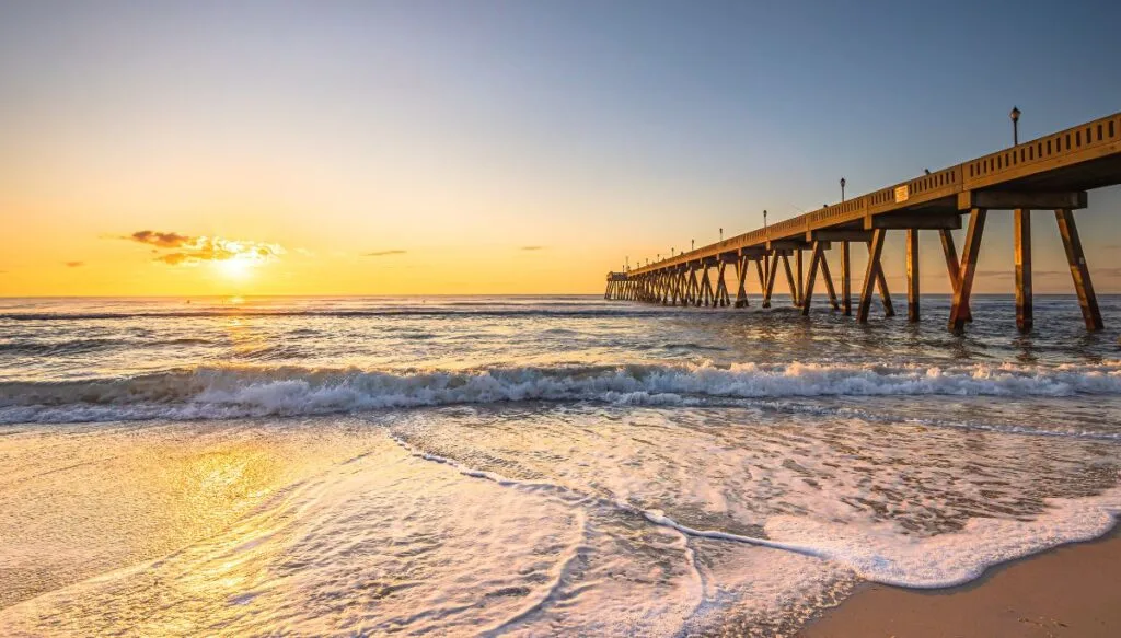 View of a wooden pier going out into the water with the sun setting in the background as the waves crash along the shore. 