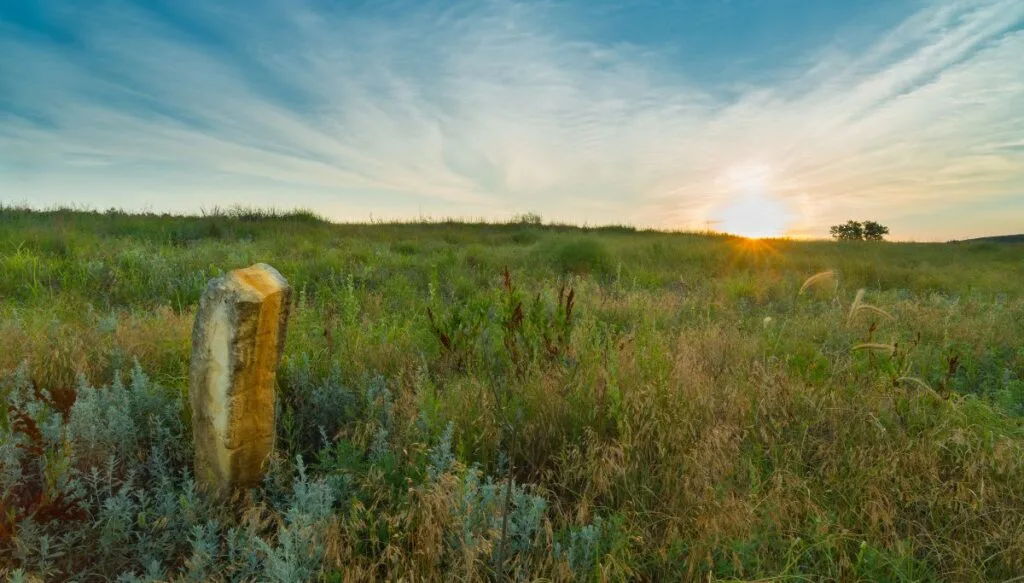 View of vast green fields with the sun setting in the background and a stone pillar in the foreground around Wilson Lake. 